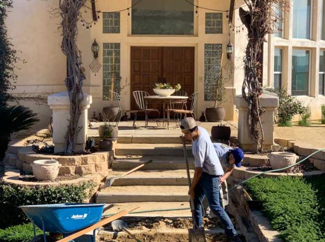 Workers renovating the front pathway of a classic home with detailed architecture, enhancing the entryway with new paving under clear skies.