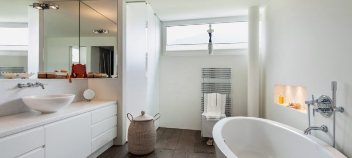 Spacious and minimalist white bathroom with a freestanding bathtub, offering a serene ocean view through large windows.
