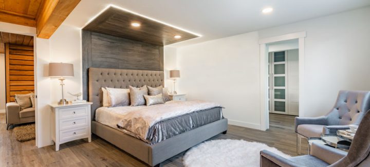 Spacious bedroom featuring a large bed with gray upholstered headboard, wooden ceiling details, and modern furnishings.